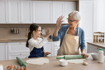 Lovely little 6s girl giving high five granny, standing together in cozy kitchen, preparing dough...