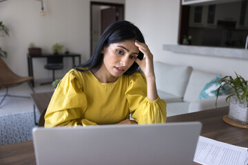 Sad concerned young Indian freelance businesswoman touching head at laptop, working on failure...