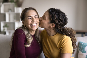 Loving young adult Brazilian daughter woman hugging and kissing happy senior Latin mom, sitting on...