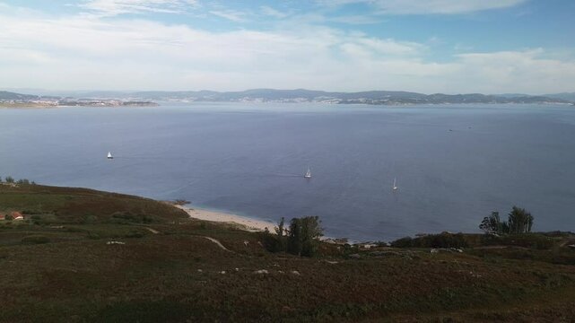 Descent over shrub-covered hillside intersected by dirt paths in Ons Island. Melide Beach with its white sand below. Three sailboats on the calm sea. Pontevedra estuary and Ald&aacute;n bay in the background