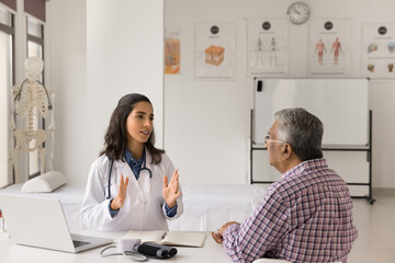 Pretty Latina female doctor in white coat provide help, consulting older male patient at his visit in private clinic. GP talk to mature man, explain treatment plan, share check-up, health test results