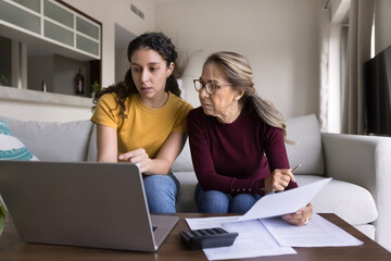 Focused Latin young daughter helping senior mother, showing how to use online financial e-bank...