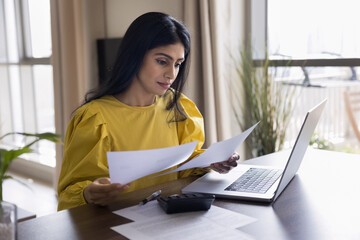 Focused Indian freelancer woman doing accounting paperwork at home, sitting at table with...