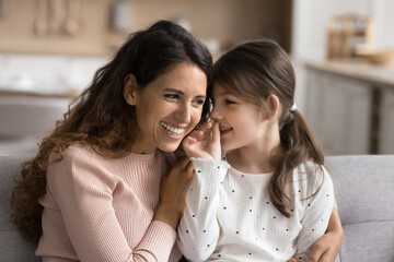 Preschooler 6s daughter sharing, whispering secrets to young loving mother, sit together on couch...