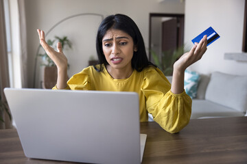 Annoyed young Indian bank client woman holding blue credit card, looking at laptop with angry...