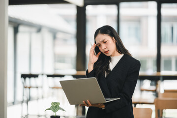 Fototapeta premium woman in black suit looks stressed while using laptop in modern office. She appears deep in thought, possibly dealing with work related challenges