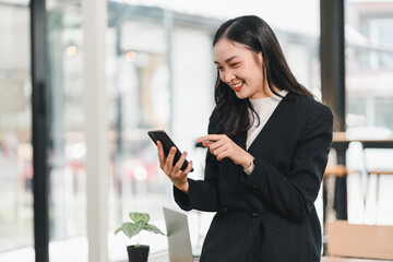 Fototapeta premium young woman in black blazer smiles while using her smartphone in modern office. Her joyful expression reflects engagement and connection