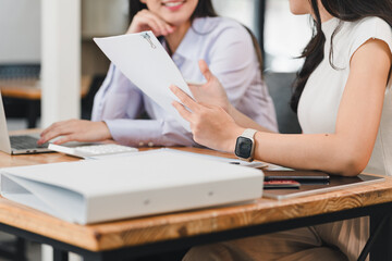Collaborative meeting between two women discussing documents at desk. One woman is holding paper while other is using laptop, creating productive work environment
