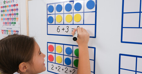 Little girl standing in classroom near white magnetic board, smiling, solving math problems using...