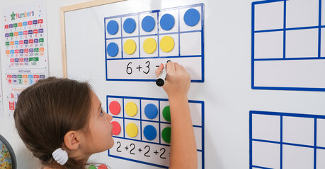 Little girl standing in classroom near white magnetic board, smiling, solving math problems using frames and counters