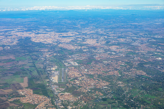 Aerial photo shows a city with dense buildings, green spaces, and a central airport. Surrounding areas include residential zones, fields, and forests. Lazio aerial view in Italy