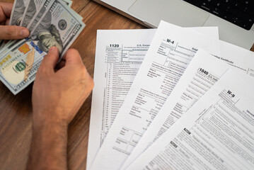 Naklejka premium Person handling cash while reviewing various tax forms on a desk. Tax preparation and financial management concept, suitable for accounting and finance themes 