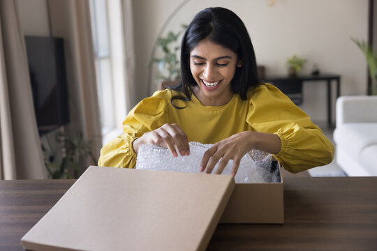 Cheerful Indian consumer woman unpacking parcel at home, opening cardboard box, unwrapping purchase from Internet store, receiving awaited package from transportation delivery company