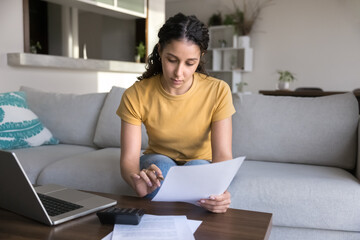 Young Latin homeowner girl reading paper invoice for payment, doing paperwork at laptop and...