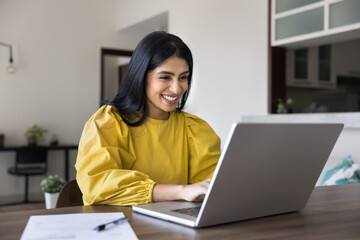 Cheerful young freelance worker girl typing on laptop at home workplace table, working at computer,...