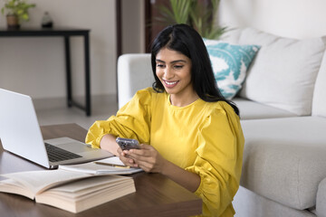 Happy young Indian student using mobile phone over open book, sitting at table with textbook and...