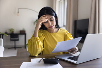 Tired concerned Indian accountant woman reading legal financial document at home workplace, holding...