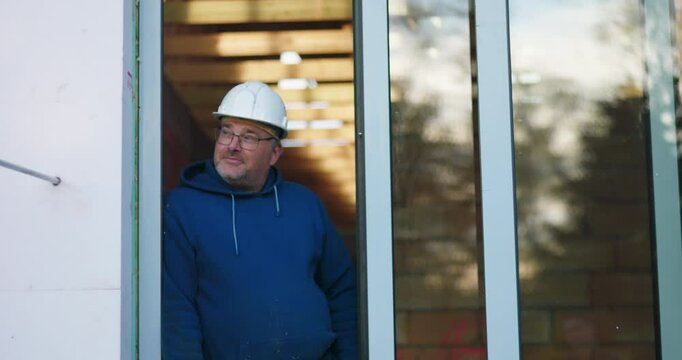 Construction worker standing in doorway, arms relaxed, wearing hard hat, observing construction progress with focus, experienced contractor in action on building site