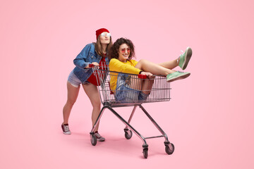 Two cheerful young friends having fun with a shopping cart in front of a vibrant pink background....
