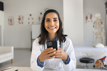Portrait of young 30s Latina doctor or nurse in white coat sit at desk with smartphone look at camera. Modern device usage in medical sphere, consulting patients remotely via mobile tele-health app