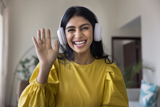 Cheerful young Indian influencer girl in wireless headphones greeting blog audience, saying hi at camera, waving hand hello, laughing, talking on video chat, giving webinar. Screen view head shot