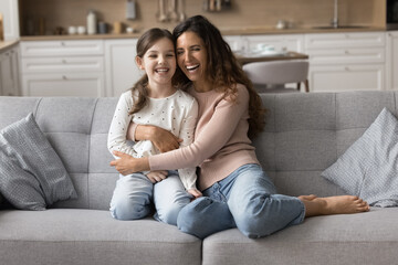 Obraz premium Laughing woman cuddling her little adorable daughter, having fun, enjoy pleasant priceless time together in cozy living room. Happy motherhood, upbringings and nurturing children, family love and ties
