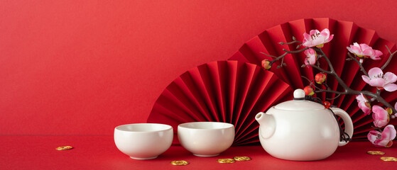 A beautiful oriental tea set displayed against a red background with cherry blossoms and a paper fan. Perfect representation of Chinese New Year 2025 tradition and festive celebrations