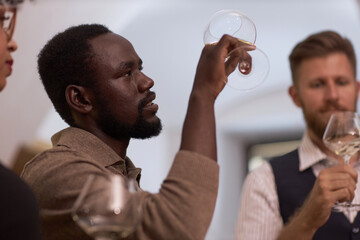 Low angle shot of African American highly skilled sommelier looking at polished crystal glass with white whine while estimating its color