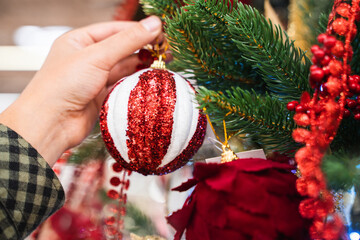 Female hand choosing a New Year Christmas red and white ball at the market, girl holding christmas tree toy decoration, Christmas decorations for sale, shopping for a classic holiday home decor