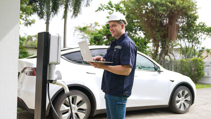 Naklejka premium Qualified technician working on home EV charging station installation, making troubleshooting and configuration setup on charging system with laptop for EV at home. Synchronos