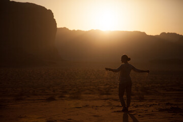 Fototapeta premium silhouette of a young beautiful girl scattering sand from her hands in the wind while being in desert during the sunset