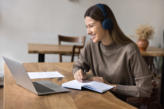 Young woman wear headphones sit at table, smiling, looking at laptop, participate in virtual communication or study session, writing in notebook, e-learns, listen audio course. Self-education, tech