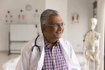 Happy Indian older man doctor smiling looking into distance standing in medical office, dressed in...