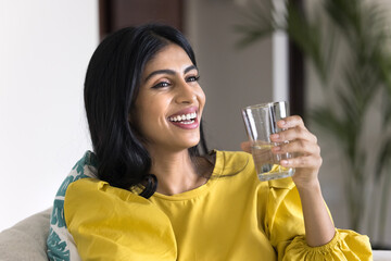 Cheerful beautiful young 20s Indian woman holding glass of pure water, drinking cold mineral beverage at home, looking away with toothy smile, laughing, enjoying healthy habit, lifestyle