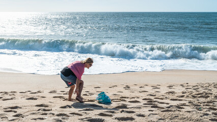 On the sandy shore of the Atlantic Ocean, a woman bends down to take off her shoes, smiling, wearing sunglasses and a red scarf; waves and white foam in the sand.