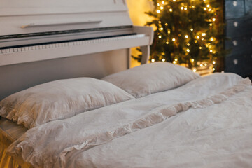 A comfortable bed featuring crisp white sheets and fluffy pillows is positioned next to a beautiful piano, accompanied by a festive Christmas tree