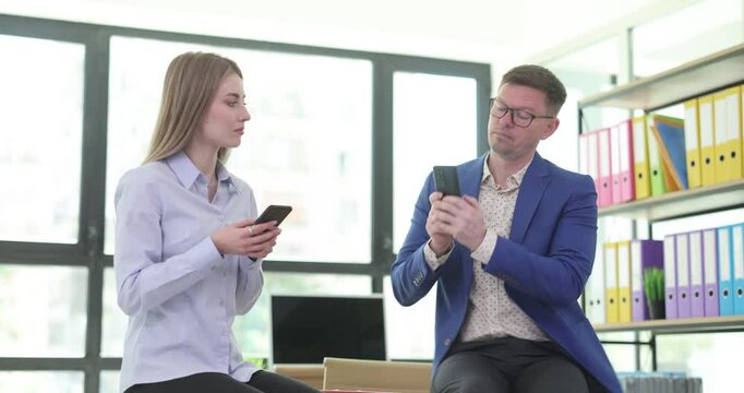 Office workers sit on table chatting in social media on smartphones. Woman tries to pry looking at guy co-worker phone and man hides screen
