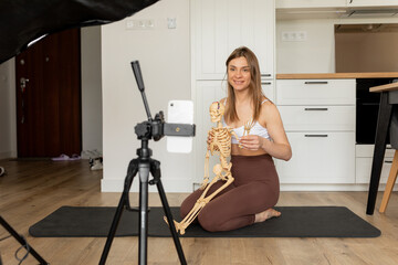 A woman is kneeling on a yoga mat, holding a skeleton and a cell phone