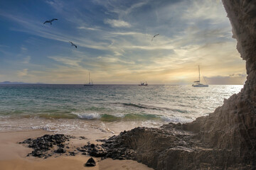 a corner of a beach next to a cliff at sunset, with boats in the background and seagulls flying...