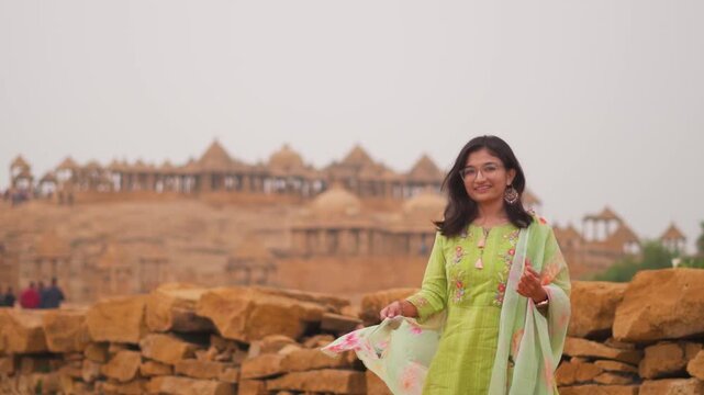 Beautiful Indian girl standing against Bada Bagh Chhatris in Jaisalmer, Rajasthan, India. Tourist exploring architecture and cultural heritage. Girl wearing traditional salwar kameez and smiling.
