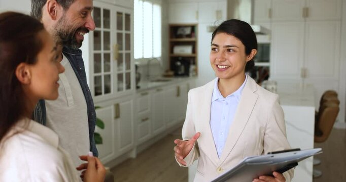 Positive young Indian realtor woman meeting with customers in home interior, holding folder of documents. Real estate, health insurance agent talking to married couple indoors