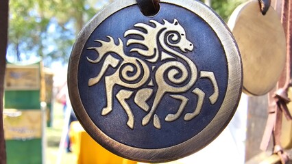 A circular medallion with an intricate golden horse design featuring Celtic-style spirals is displayed outdoors, with blurred greenery and market-like structures in the background.