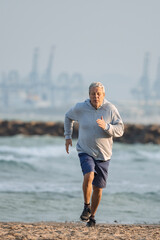 Caucasian man in grey hoodie and blue shorts running along a sandy beach with industrial structures visible in the distance