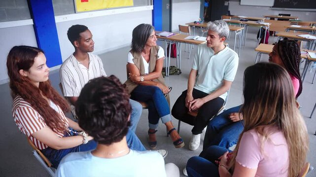 Diverse happy group of high school students sitting on chairs in circle interacting during lesson. Multiracial adolescents talking together to their classmates and female teacher in support class