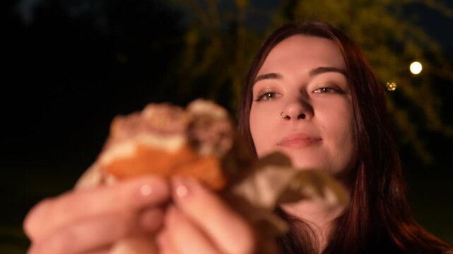 Closeup view of female Caucasian person biting cheeseburger and chewing it with satisfaction on her face sitting in the street at night time. Cheerful lady is enjoying delicious fast food outdoors