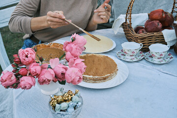Woman delicately spreads caramel on waffles at an outdoor table adorned with pink peonies and apples. A charming scene of homemade treats and rustic elegance.