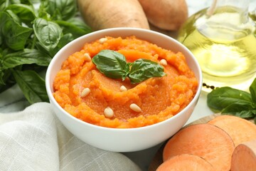 Tasty mashed sweet potato with basil, seeds in bowl and ingredients on white table, closeup
