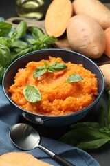 Tasty mashed sweet potato with basil in bowl, fresh vegetables and spoon on table, closeup
