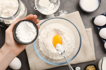 Woman adding baking powder into bowl at grey table with other products, top view