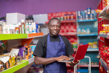 A grocery store employee in an apron uses a laptop for inventory management. Shelves are stocked with various products in a colorful retail environment.